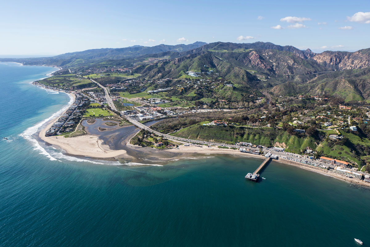 Wide aerial view of Malibu, California, featuring the Pacific coastline, Malibu Pier, surrounding beaches, and the Santa Monica Mountains.